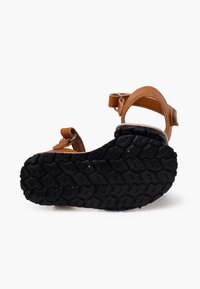 Sandalo in pelle marrone con fasce regolabili, suola in gomma testurizzata e un motivo di battistrada unico. Presenta un design aperto sul retro per una migliore traspirabilità.
