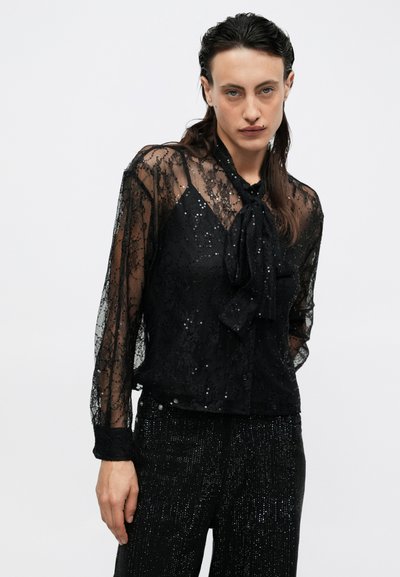 Sort bluse i blonde med en bindesnor ved halsen, der har pailletdetaljer og gennemsigtige lange ærmer. Strukturstoffet tilføjer et lagdelt udseende.