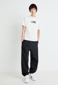 T-shirt in cotone bianco con stampa grafica nera, abbinato a pantaloni felpati neri a sigaretta con polsini elasticizzati e tasche laterali, indossato con sneakers bianche.