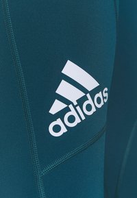 Sportovní legíny v tyrkysové barvě s výrazným bílým logem Adidas. Vyrobeny z hladkého, pružného materiálu s plochými švy pro pohodlí.