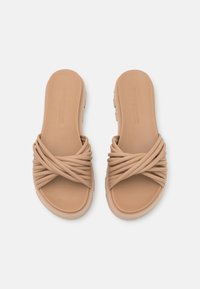 Sandales à glissière en caoutchouc beige avec un design de sangle texturée et torsadée. Dispose d'une semelle plate et d'un coussin de pied lisse.