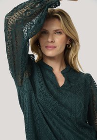 Blusa in pizzo di un blu scuro con motivi geometrici, maniche lunghe trasparenti e scollo a V. Presenta cuciture intricate e dettagli testurizzati.