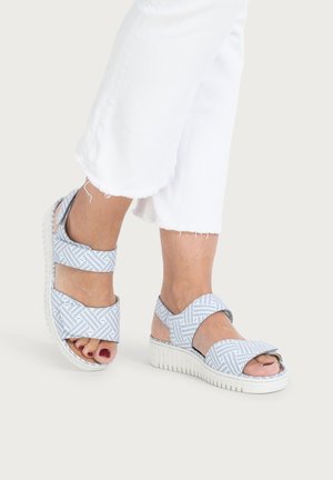 Füße, die hellblau-weiß gemusterte Sandalen mit offenen Zehen und verstellbaren Riemen tragen, kombiniert mit weißen, gekürzten, ausgefransten Hosen.