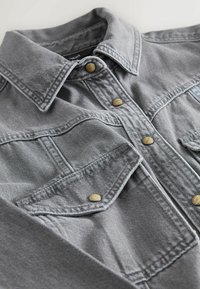 Graue Jeansjacke mit Kragen, zwei Brusttaschen und goldenen Druckknöpfen. Sie hat einen strukturierten Stoff und sichtbare Nähte.