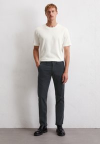 Weißes kurzärmliges T-Shirt, kombiniert mit dunkelgrauen maßgeschneiderten Hosen und schwarzen Schnürschuhen, vor einer strukturierten hellen Wand stehend.