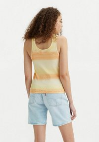 Vrouw met krullend haar, gekleed in een mouwloze oranje en gele gestreepte tanktop en lichtblauwe denimshorts, met de rug naar ons gekeerd.