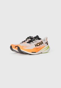 Chaussures de course avec une tige en maille présentant des accents orange et noir. Semelle épaisse blanche avec un design vert et orange. Logo HOKA sur le côté.