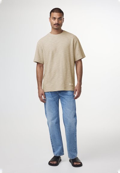 Camicia beige a maniche corte con texture e collo a giro, abbinata a jeans dritti azzurri e sandali neri con zeppa.