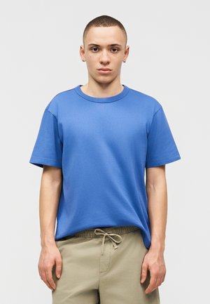 Νεαρός άνδρας με κοντά μαλλιά φορώντας μια μονόχρωμη μπλε κοντομάνικη μπλούζα και μπεζ παντελόνι με κορδόνι στη μέση, μπροστά σε ανοιχτό γκρι φόντο.