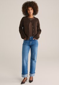 Hnědý chlupatý cardigan s kulatým výstřihem a zapínáním na knoflíky, nošený přes hnědý top a doplněný modrými džíny s ohrnutými manžetami.