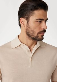 Bežs īsām piedurknēm polo krekls ar teksturētu audumu, trīs pogu aizdari un kontrastējošu apkakli. Parādīts modeļa sānu profila skatījumā.