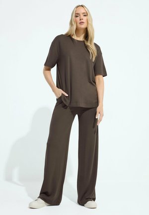 Donna bionda in piedi, indossa una maglietta a maniche corte larga di colore marrone scuro e pantaloni a gamba larga con sneakers bianche su sfondo bianco uniforme.