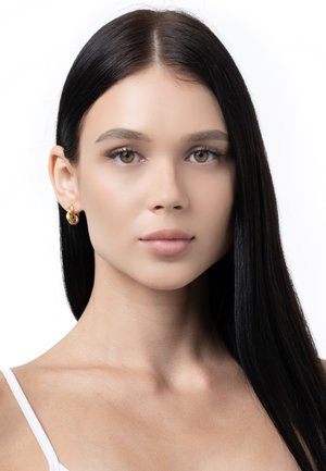Goldene Creolen zeigen ein geschichtetes, ineinander verwobenes Design. Das Modell hat glattes, dunkles Haar und einen neutralen Gesichtsausdruck vor einem weißen Hintergrund.