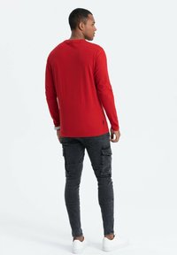 Rode langelijfshirt met ronde hals, gecombineerd met donkergrijze cargo-broeken. Model met witte sneakers, met de rug naar de camera gekeerd.