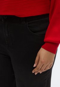 Top roșu cu mâneci lungi și dungi, asortat cu blugi negri cu talie înaltă, având un buzunar frontal și textură fină. Mână sprijinindu-se pe blugi.