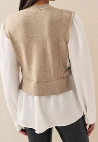 Veste beige en tricot avec une texture côtelée, taille ajustée et ceinture, portée sur un chemisier blanc et un pantalon noir. Vue de dos.