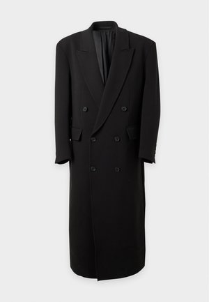 Homme portant un long manteau noir, un pantalon noir, des chaussures noires, un pull à col roulé blanc, une ceinture noire et un bonnet brun foncé, debout avec une main dans la poche.