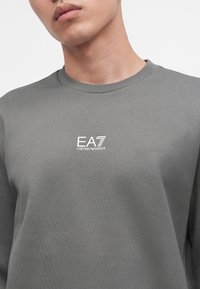 Gros plan d'une personne portant un t-shirt gris à col rond avec le logo blanc "EA7 Emporio Armani" centré sur la poitrine.