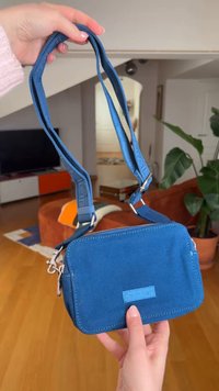 Χέρια που κρατούν μια μικρή μπλε τσάντα Calvin Klein με ρυθμιζόμενο λουρί σε ένα ζεστό καθιστικό.