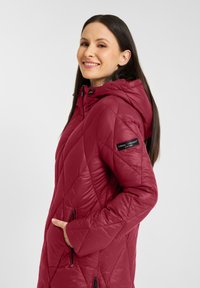 Vrouw met een glimlach, gekleed in een rode gewatteerde jas met capuchon en haar hand in de ritszak aan de zijkant, zichtbaar merkembleem op de linkermouw.