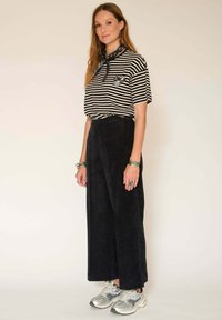 Chemise rayée noire et blanche, à manches courtes, avec une poche avant ; pantalon large en velours côtelé noir ; baskets argentées avec accents noirs ; bracelets superposés.