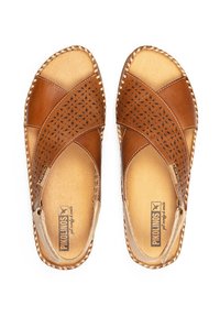 Sandalias de cuero marrón con un diseño perforado, bordes cosidos y suela plana. El interior cuenta con un forro en tono beige y una marca.