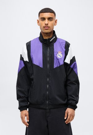 Ein junger Mann trägt eine schwarze, violette und weiße Adidas-Trainingsjacke mit einem Real-Madrid-Logo auf der Brust und steht vor einem schlichten Hintergrund.
