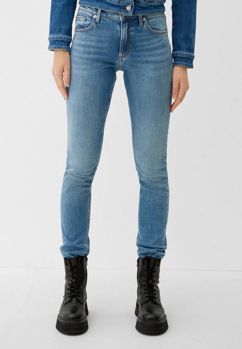 Femme portant un jean skinny bleu clair, une veste en jean et des bottes de combat noires à lacets, debout devant un fond uni.