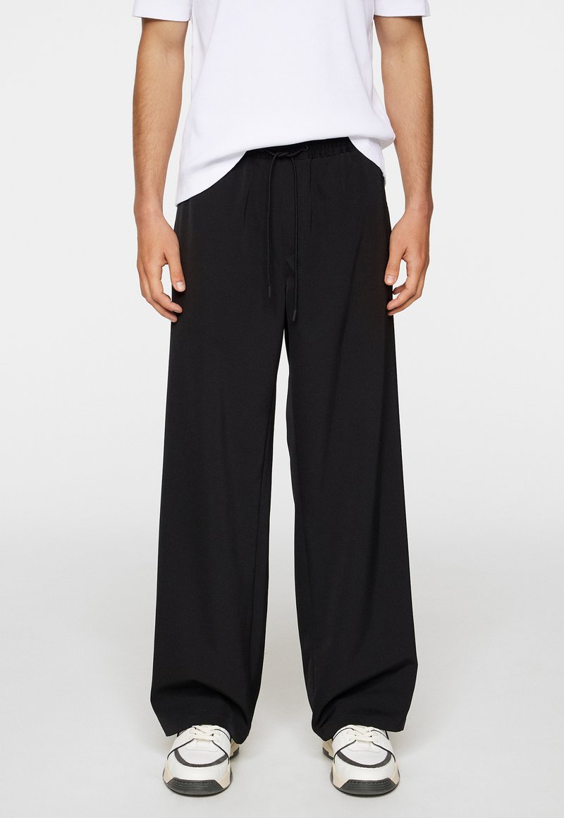 Black loose-fitting pants with an elastic waistband and drawstring. Smooth fabric with a relaxed silhouette, paired with white sneakers.