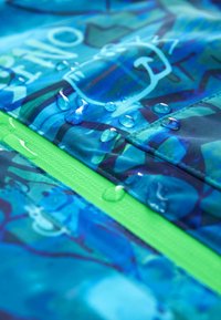 Gros plan de gouttes d'eau perlant sur un tissu imperméable bleu et vert avec des motifs graphiques et des détails de couture.