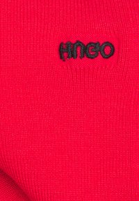 Czerwony dzianinowy materiał o teksturowanej powierzchni z małym czarnym haftowanym logo "HUGO" umieszczonym centralnie.