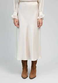 Une jupe midi crème, en tissu lisse, présente une coupe évasée. Associée à des bottes de cowboy en daim marron avec un piquage élaboré.