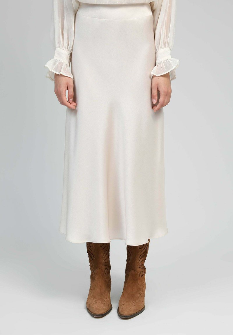 Une jupe midi crème, en tissu lisse, présente une coupe évasée. Associée à des bottes de cowboy en daim marron avec un piquage élaboré.