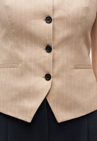 Gilet beige à fines rayures avec trois boutons noirs centrés et deux poches horizontales, porté sur un pantalon noir.