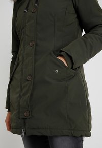 Personne portant un manteau matelassé vert foncé avec des boutons et une fermeture éclair, une main dans la poche avant, associée à un jean foncé.