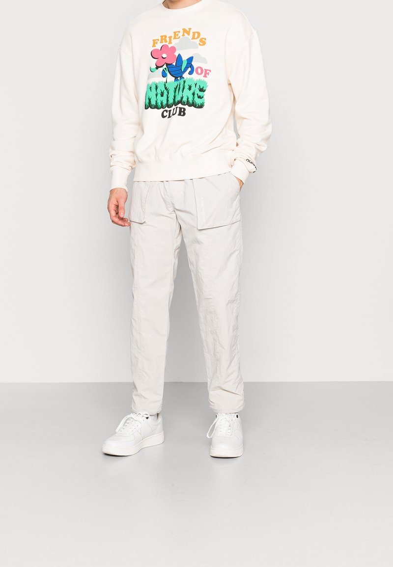 Persona vestida con un sudadera crema con un gráfico de "Club de Amigos de la Naturaleza", pantalones cargo color beige claro y zapatillas blancas, de pie contra un fondo liso.