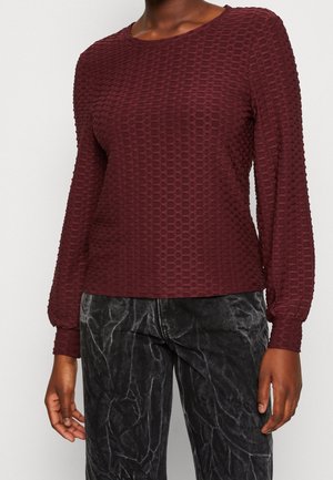 Femme portant un haut bordeaux texturé à manches longues associé à un pantalon en velours noir à l'aspect froissé.