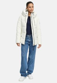 Vrouw in een witte gewatteerde jas met capuchon, marineblauwe top, losse blauwe spijkerbroek en witte sneakers, staand tegen een effen achtergrond.