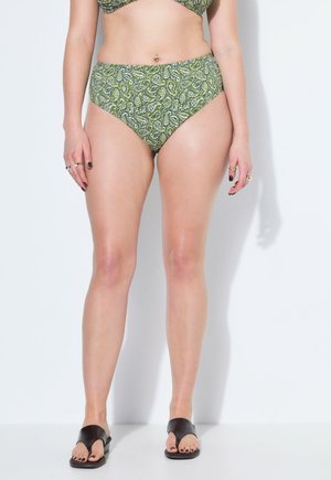 Kobieta w zielonych, wysokich majtkach bikini w wzór paisley oraz czarnych klapkach stoi na tle białej ściany.