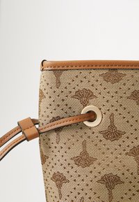 Vaaleanruskea käsilaukku, jossa on rei'itetty kangas ja ruskeat kukkakuvioinnit. Siinä on nahkareunus ja kullanvärinen nitoja.