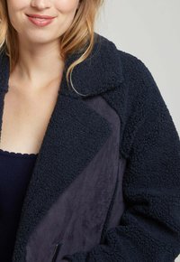 Veste bleu marine en tissu doux avec un extérieur pelucheux et un intérieur lisse. Dotée d'un large col et d'une fermeture éclair.