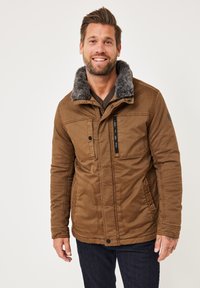 Brown, textured jacket with a faux fur collar, multiple pockets, and zippered detailing. Worn over a knit sweater and jeans.