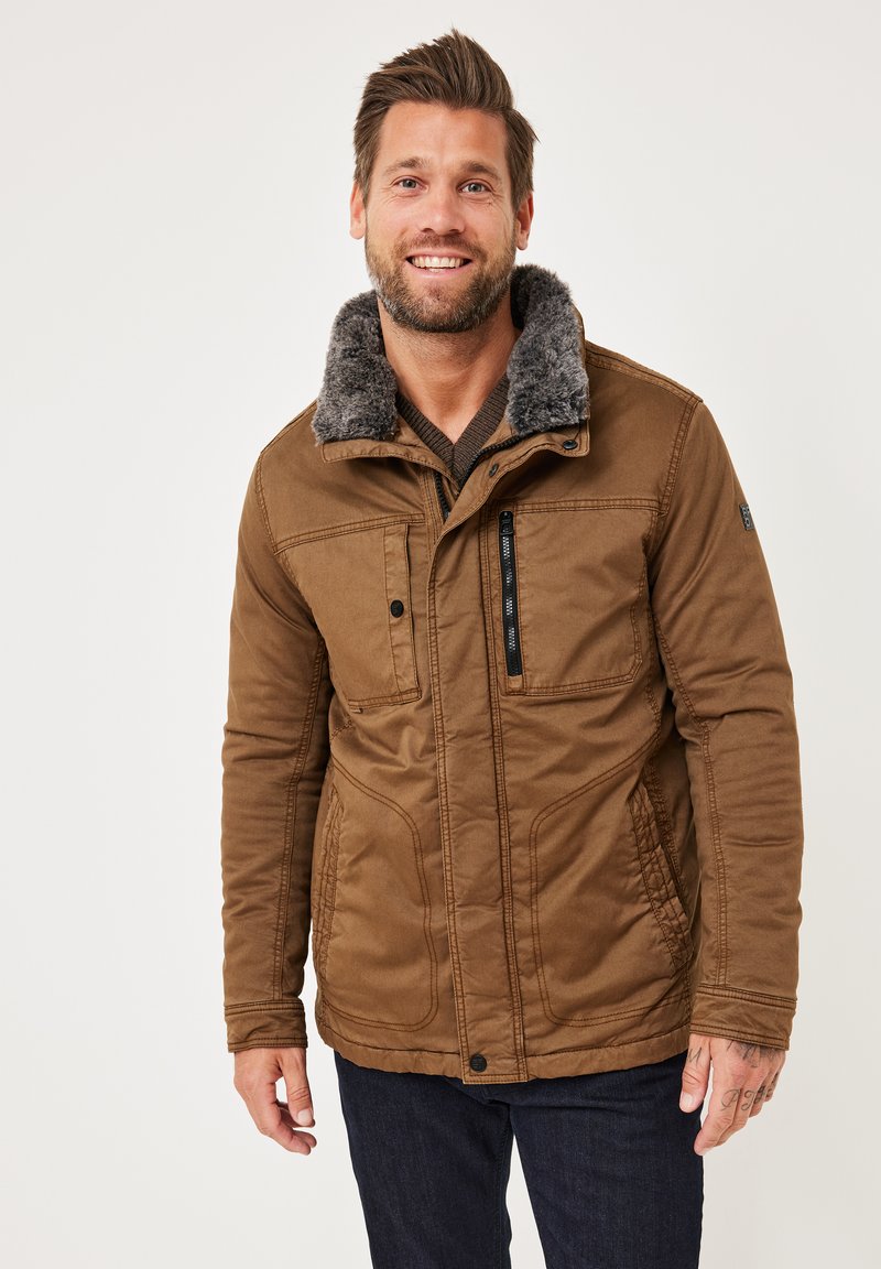 Brown, textured jacket with a faux fur collar, multiple pockets, and zippered detailing. Worn over a knit sweater and jeans.