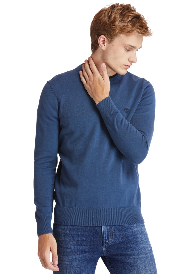 Maglione blu a maglia con scollo rotondo, polsini e orlo a costine. Presenta un piccolo logo sul petto. Indossato con jeans blu.