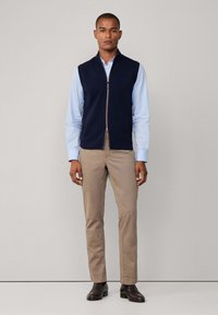 Tamno plavi bezrukavnik s patentnim zatvaračem, nosi se preko svijetloplavog dugih rukava; uparen s bež hlačama i tamnim cipelama.