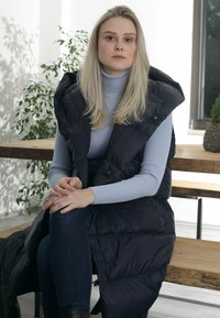 Langer, dunkler Daunenwesten mit hohem Kragen, getragen über einem hellblauen, gerippten Rollkragenpullover. Dazu passen Jeans und schwarze Stiefeletten.