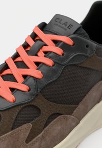 Sneaker con un mix di suede marrone scuro e rete testurizzata, con lacci arancione brillante e un accento in pelle nera vicino alla linguetta.