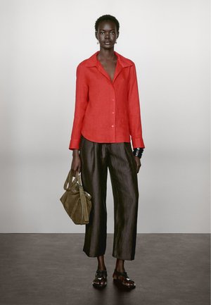 Red linen button-up shirt with a collar, paired with dark, wide-leg pants. Model holds a green suede bag and wears black sandals.