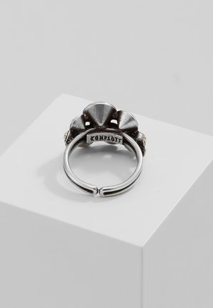 Verstelbare zilverkleurige ring met drie getextureerde, driehoekige metalen accenten en een open band, liggend op een wit kubusvormig voetstuk.