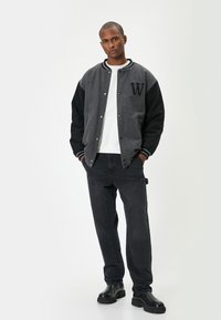 Chaqueta varsity gris y negra con un parche de "W", puños acanalados, botones a presión, combinada con pantalones negros y botas negras gruesas.